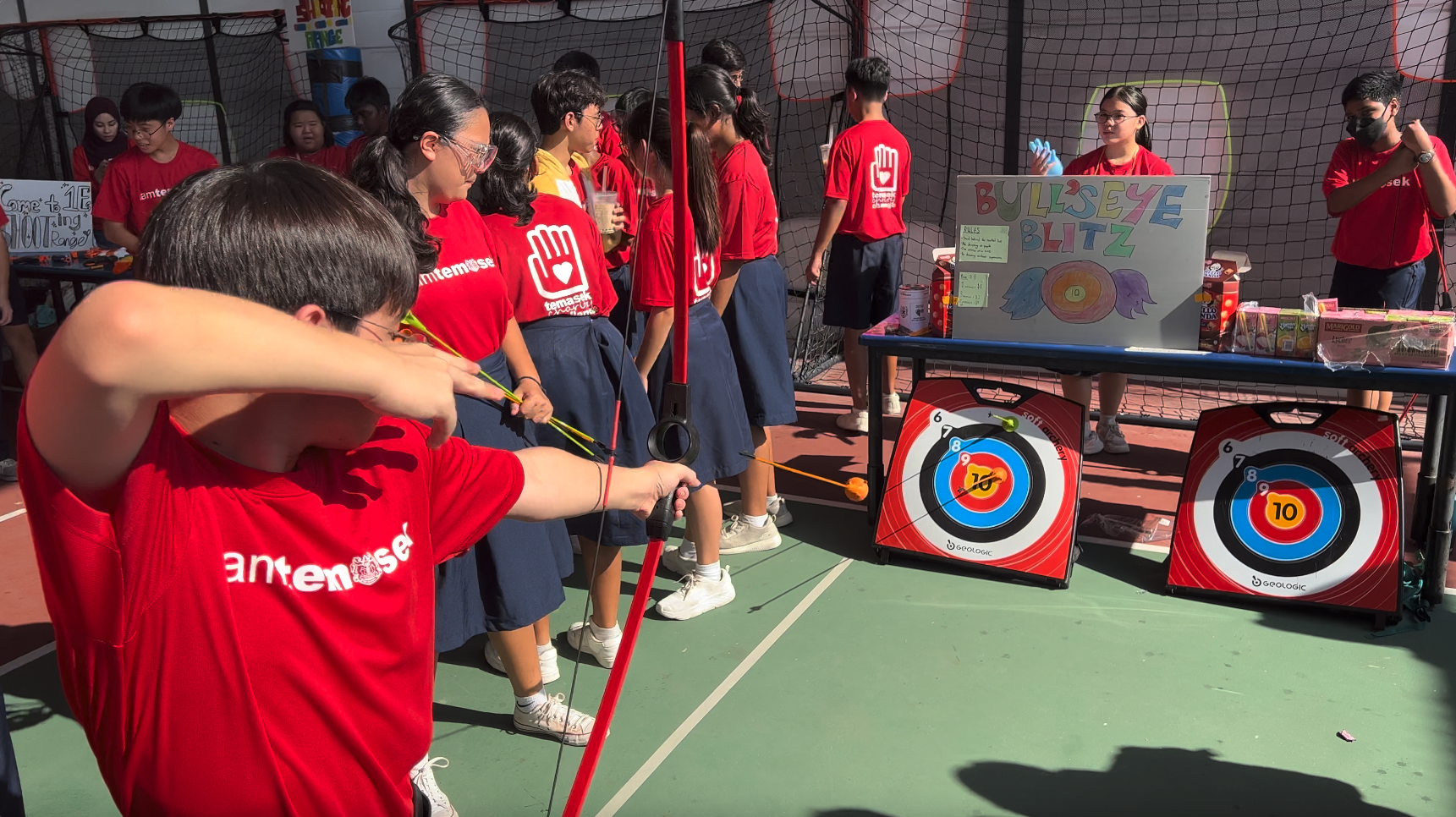 Students in red shirts play a Bulls Eye Blitz archery game outdoors, with a boy aiming a bow at circular targets.